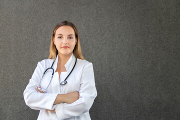 Portrait of modern female doctor posing in studio on gray background. Woman wearing a laboratory coat, holding stethoscope, crosses her arms. Copy space. Medicine, healthcare and profession concept.