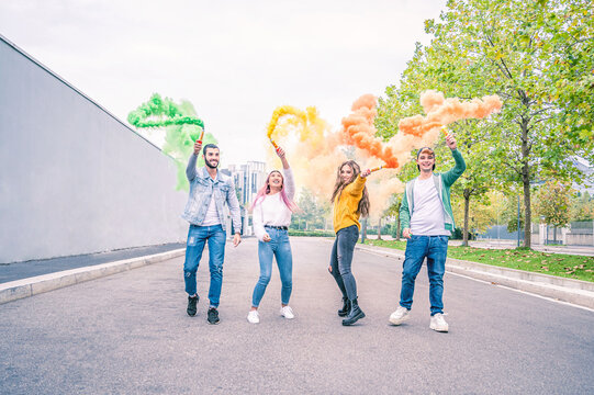 Happy smiling young people running at street with a coloured signal rocket in the hand - Happy friendship concept with young students having fun together outdoors with smoke flare