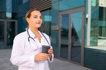 Energetic charge before beginning of working day of female therapist standing at entrance to hospital, wearing in white lab coat, holding a mug of tea. Copy space. Medicine and healthcare concept.
