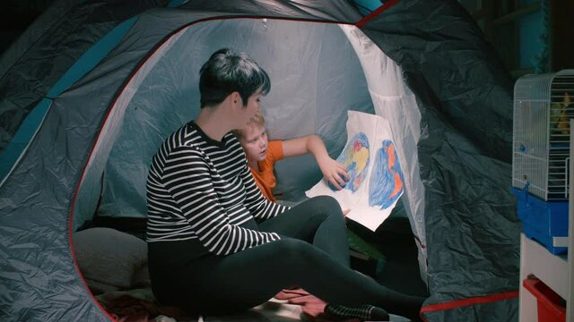 A pregnant mother with her son sitting in a tent looking at a homemade map of the world. Home education in self-isolation.