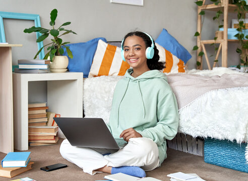 Happy mixed race teen school girl distant college student wearing headphones virtual remote e learning using laptop in bedroom looking at camera. Distance education classes, studying online at home.