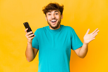 Young indian man holding a phone receiving a pleasant surprise, excited and raising hands.