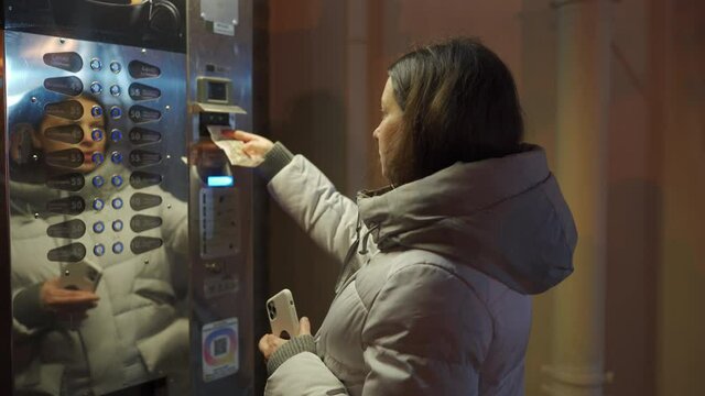 Woman buying coffee from vending machine and pushing some buttons on an empty street at cold night. Pavlovsk Park and Palace in autumn, St. Petersburg, Russia.