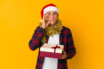 Young caucasian man with christmas hat holding a present isolated on yellow background