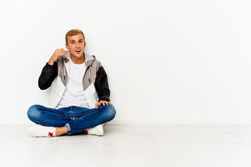 Young caucasian man sitting on the floor laughing about something, covering mouth with hands.