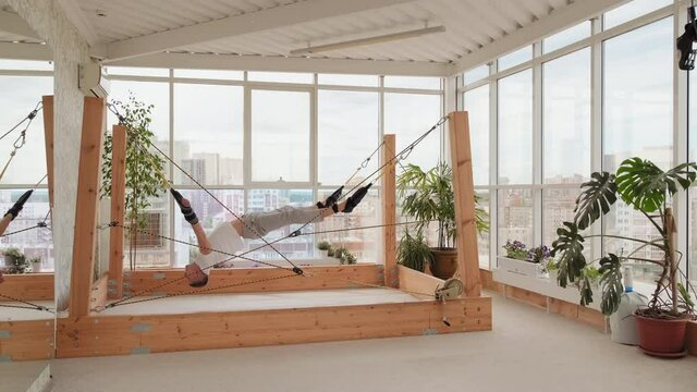 Full shot of man attached by ropes to fasteners parallel to floor twisting in air stretching body and strengthening ligaments on futuristic simulator