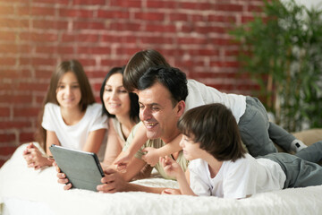Relaxing together. Joyful latin family with cute little kids spending time together at home. Father using digital tablet, lying on the bed with children