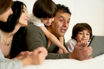 Stay connected. Happy latin family with little kids using digital tablet, lying on the bed together. Parents and children looking at the screen while having video call in the morning