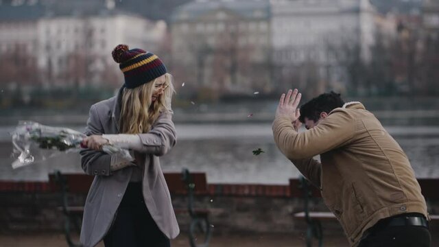 Crazy caucasian girl taking bouquet of flowers and brutally hiting guy on river embankment. Young couple screaming and quarreling on date. Jealous woman beating scared man with bouquet after betrayal.