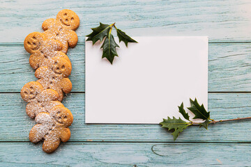 gingerbread cookies, gingerman, arranged on a blue table with holly, traditional Christmas plant, and a blank white card with copyspace, lets you create a tasty Christmas card - 396663006