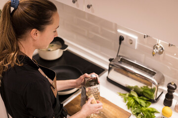 Young beautiful woman standing by the stove and grating the cheese for a healthy food recipe....