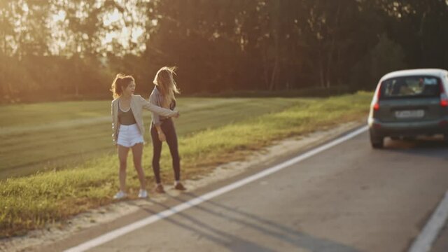 Pretty women travelers waiting for car s at roadside. Young sexy girls dressed in casual summer clothes hitchhiking on the road raising their thumbs up. Concept of freedom and travelling.