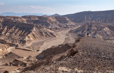Fototapeta na wymiar Young female on a trail in remote region of Eilat mountains, Israel. Desert beauty in a sunny winter day. Panoramic landscape of colorful sandy hills, dry wadies, rock formations, mountains, boulders.