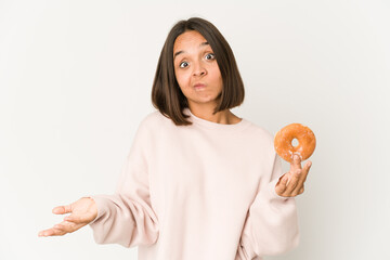 Young hispanic woman eating a doughnut shrugs shoulders and open eyes confused.