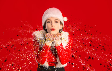 Sweet  girl wearing Santa Claus costume blowing sparkle magic dust, on red background, holiday...