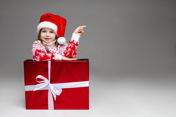 Stock photo of sweet little girl in patterned winter sweater and Santa hat indicating at blank space leaning on wrapped Christmas present with white bow. Isolate on grey background.
