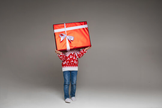 Stock photo of anonymous kid in jeans and winter sweater carrying giant Christmas gift wrapped in red paper with white bow. Cutout on grey background. Studio shot.