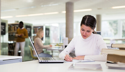 Fototapeta premium Adult female student with laptop and books in public library. High quality photo