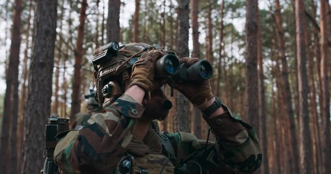 During a reconnaissance mission, the Squad Leader of fully equipped camouflaged soldiers looks for the enemy through binoculars in a thick pine forest.