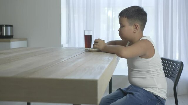 Overweight Asian boy eating unhealthy burger and drinking cola at home
