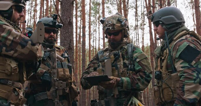 A group of middle-aged bearded soldiers in uniforms and tactical vests discusses the action plan and prepares for action in a dense forest. The commander gives instructions to the soldiers.