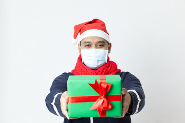 A portrait of a young asia man wearing a face mask and a Santa Claus hat standing in front of a white background and holding a Christmas gift box