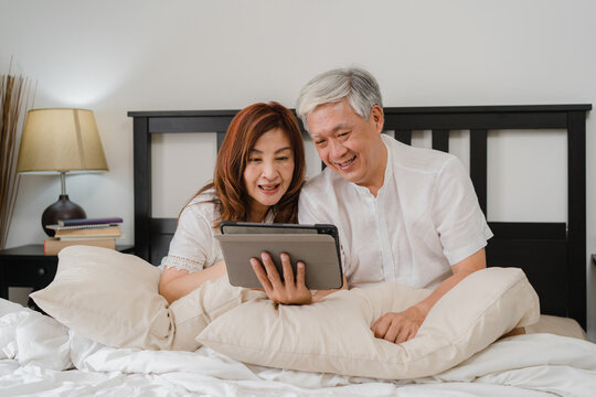 Asian senior couple using tablet at home. Asian Senior Chinese grandparents, husband and wife happy after wake up, watching movie lying on bed in bedroom at home in the morning concept.