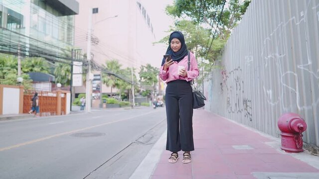 Attractive female asian muslim holding takeaway coffee cup and head down scrolling on smartphone while standing on a footpath next busy street, taxi calling application, convenience technology user