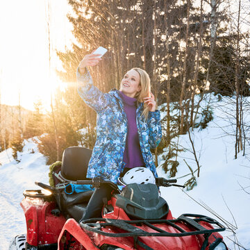 Joyful blonde woman in winter jacket taking picture with smartphone during quad biking in winter mountains. Charming female traveler on all-terrain vehicle looking at phone camera and smiling.