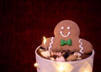 Delicious cup of hot chocolate inside a white cup with a gingerbread man cookie dipped in it with chocolates on top sprinkled with cinnamon on a red background and Christmas lights