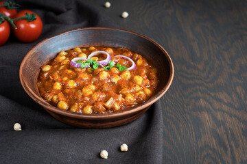 Chole masala or chana indian food made of cooked chickpeas, tomatoes and cumin decorated with onion rings and parsley served in bowl on textile on dark brown wooden background. Image with copy space