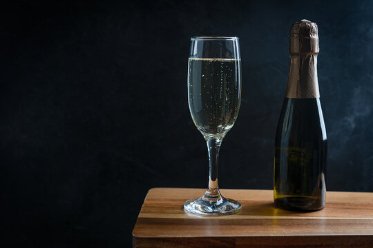 Cold white Prosecco sparkling champagne served in tall drinking flute glass with small bottle on dark brown wooden bar counter at party against black background. Image with copy space, horizontal