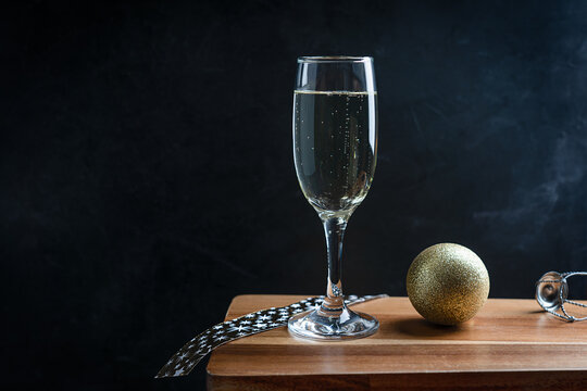 Cold white Prosecco sparkling champagne served in tall drinking flute glass with golden christmas ball, ribbon and wire hood on dark wooden bar counter against black background. Image with copy space