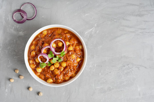 Top view of chole masala or chana indian food made of cooked chickpeas, tomatoes and cumin decorated with onion rings and parsley served in bowl on gray concrete background. Image with copy space