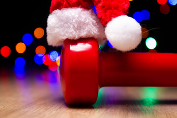 Heavy red dumbbell with Santa Claus hat. Exercise equipment as a Christmas gift idea. Healthy fitness lifestyle holiday season concept. Composition with colorful bokeh lights in background.