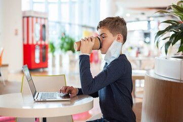 Distance learning. A teenage boy in a medical mask sits at a table in a cafe with a laptop and a mug of hot drink.