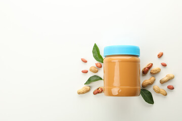 Jar with peanut butter, peanuts and leaves on white background