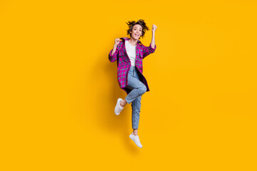 Fototapeta na wymiar Full size photo of triumphant happy girl wear jeans violet checkered cardigan raise fists jump air isolated on yellow color background