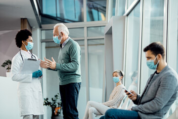 Senior man and African American doctor wearing face masks while talking in the hospital.