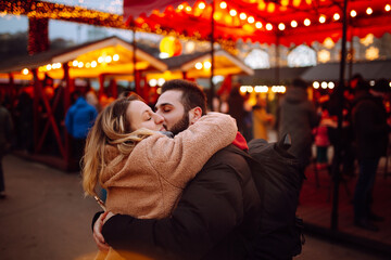Obraz premium Young couple outdoors at winter on a new year`s eve having fun at a winter fairy, lights all around, souvenir shops. Happy woman and man visits the decorated Christmas market during the evening.