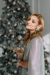 Smiling beautiful blonde woman in a luxurious outfit with a glass in her hands against the background of blurry lights bokeh of a Christmas tree. Happy New Year 2021. Soft selective focus, art noise.