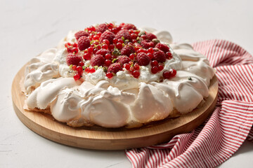 food, culinary, baking and cooking concept - close up of pavlova meringue cake decorated with red berries on wooden serving board and kitchen towel