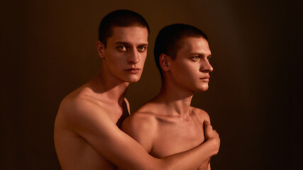 Two young half naked caucasian twin brothers posing together in studio, standing isolated over brown background
