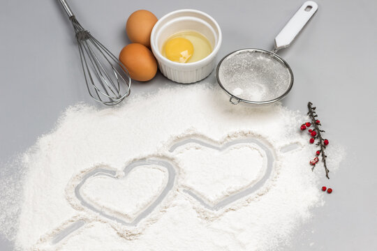 Two hearts are painted on flour. Broken egg in bowl, whisk and sieve on table. Ingredients for baking cookies