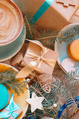 Top view on Christmas gifts with Christmas decorations on a wooden table with blue cup with cappuccino and sweet macarons.
