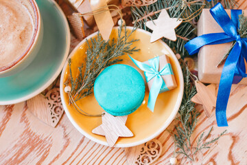 Top view on yellow saucer with blue macaron, with Christmas gift, wooden decorations, coniferous branches and blue cup with cappuccino on a wooden background.