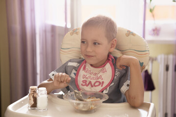 Fototapeta premium a boy with a bib eats porridge at the children's table