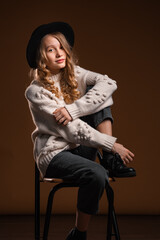 Charming blonde teen girl in black hat posing in studio on brown background