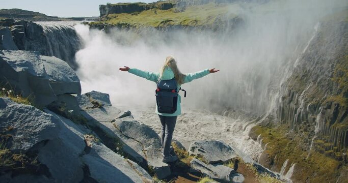 Woman standing on the edge of the cliff and raising her hands up in the air wiht beautiful Detifoss waterfall in background. Iceland