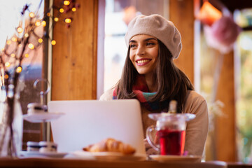 Smiling young woman in smart casual wear sitting behind her laptop at the cafe while looking thoughtfully.  